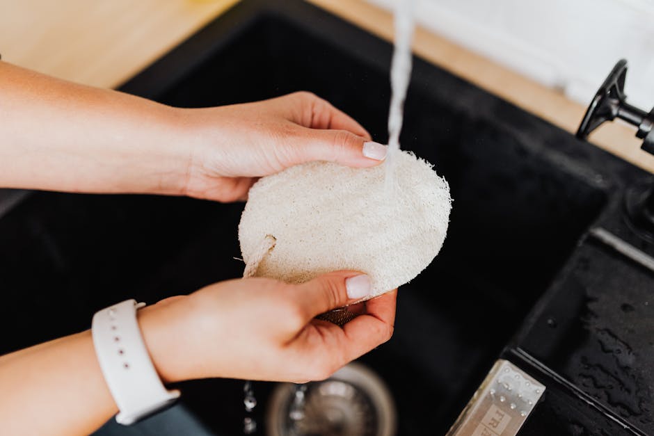 A woman rinses a loofah under running water in a kitchen sink. Hygiene and care concept.